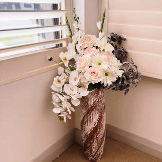 Blush pink and white artificial flower arrangement in a tall textured vase beside a window