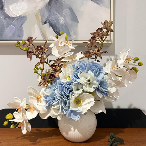 Customer photo of blue and white flowers with white orchids and brown orchid stems in a white vase.